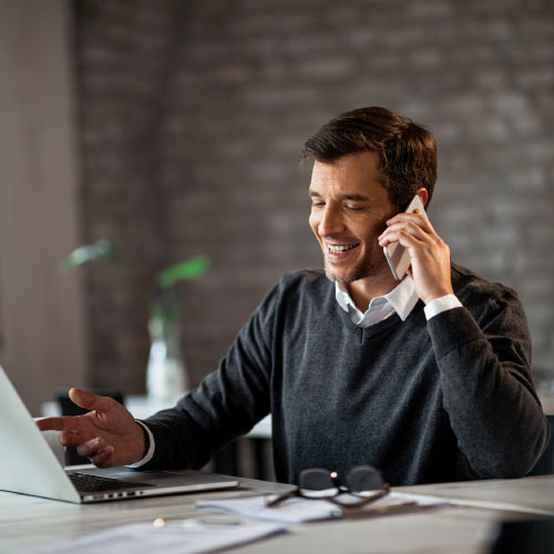 A smiling man in a smart cardigan sits at a laptop while on the phone.