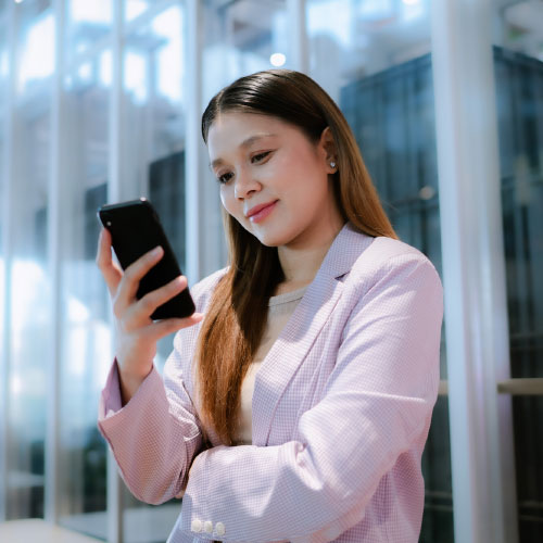 An asian businesswoman dressed in a pink blazer scrolls through her smartphone while smiling